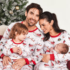 Portrait of a smiling mother, father, toddler, and newborn baby cuddling together in matching Santa and sleigh pattern pajamas next to a Christmas tree.
