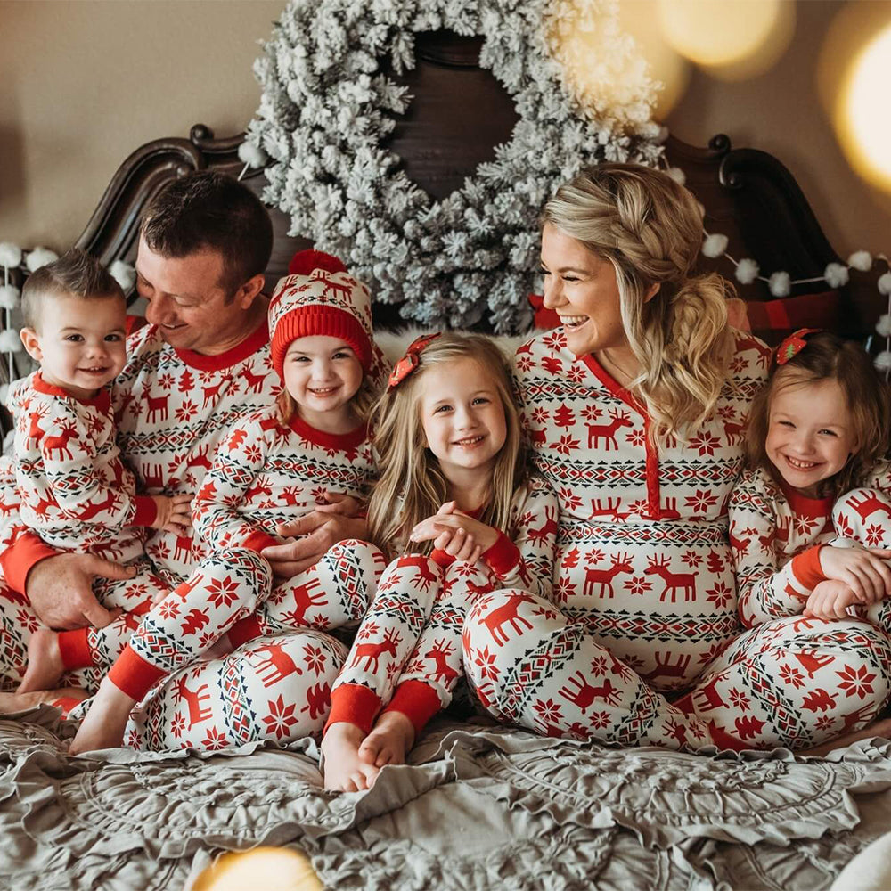 Parents and four young children cuddling together on a decorated bed wearing matching red and white Fair Isle reindeer Christmas pajamas.