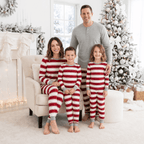Family in red and white striped pajamas around an armchair by a snowy Christmas tree, dad in grey.