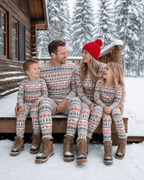 Family in grey Fair Isle Christmas pajamas with red and green motifs sitting on snowy cabin steps.