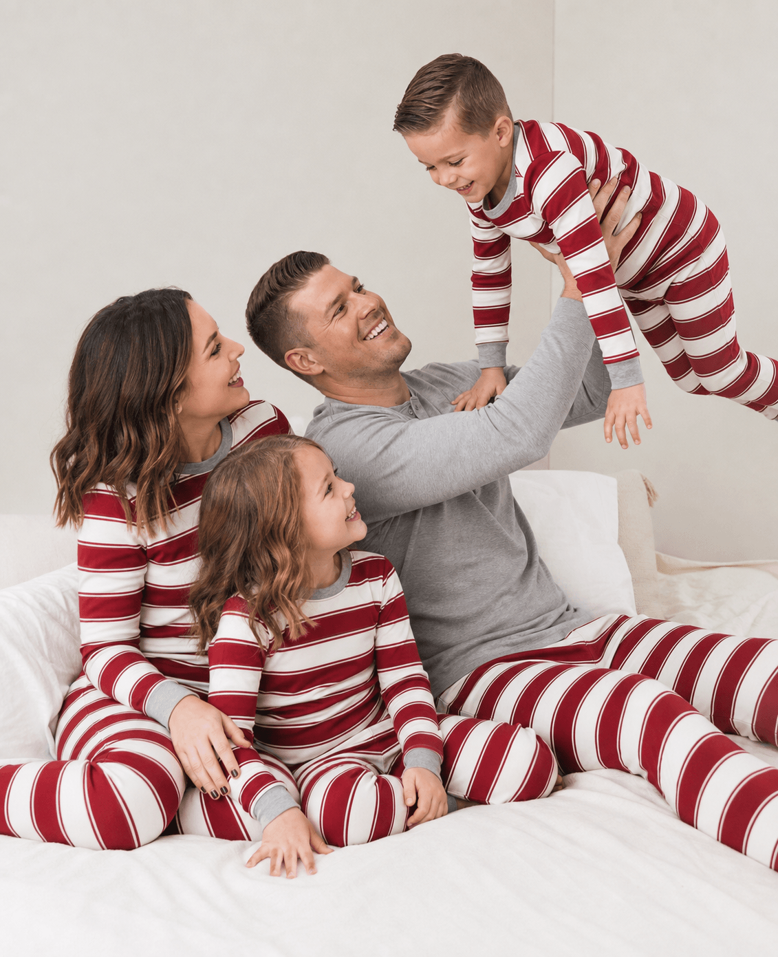 Dad lifting his son above a bed as the family plays in red and white striped pajamas with grey trim.