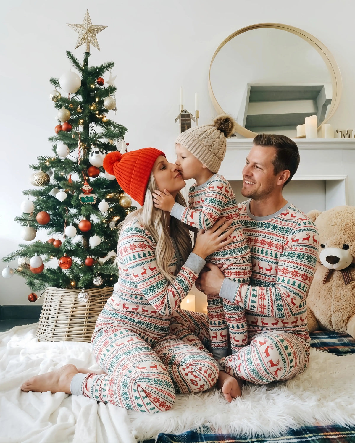 Mother kissing her child beside a Christmas tree as dad watches, all wearing matching grey Fair Isle pajamas.