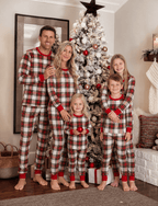 Parents and three children in matching red and white plaid Christmas pajamas beside a snowy tree.