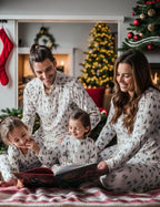 A family of four reading a holiday storybook together on the floor in matching white pajama sets featuring a green watercolor pine tree print next to a decorated Christmas tree.
