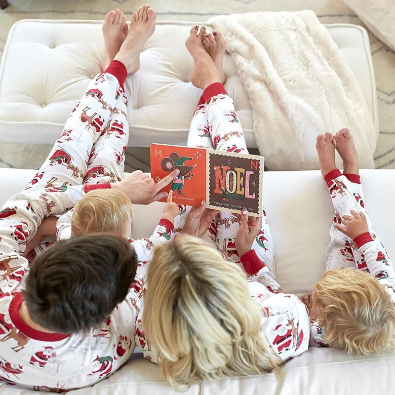 Top-down view of parents and two kids lying on a sofa reading a NOEL holiday book together in matching Santa and reindeer pajamas.