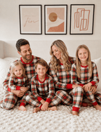 Parents and three children smiling together on a bed in red and white plaid Christmas pajamas with red cuffs.