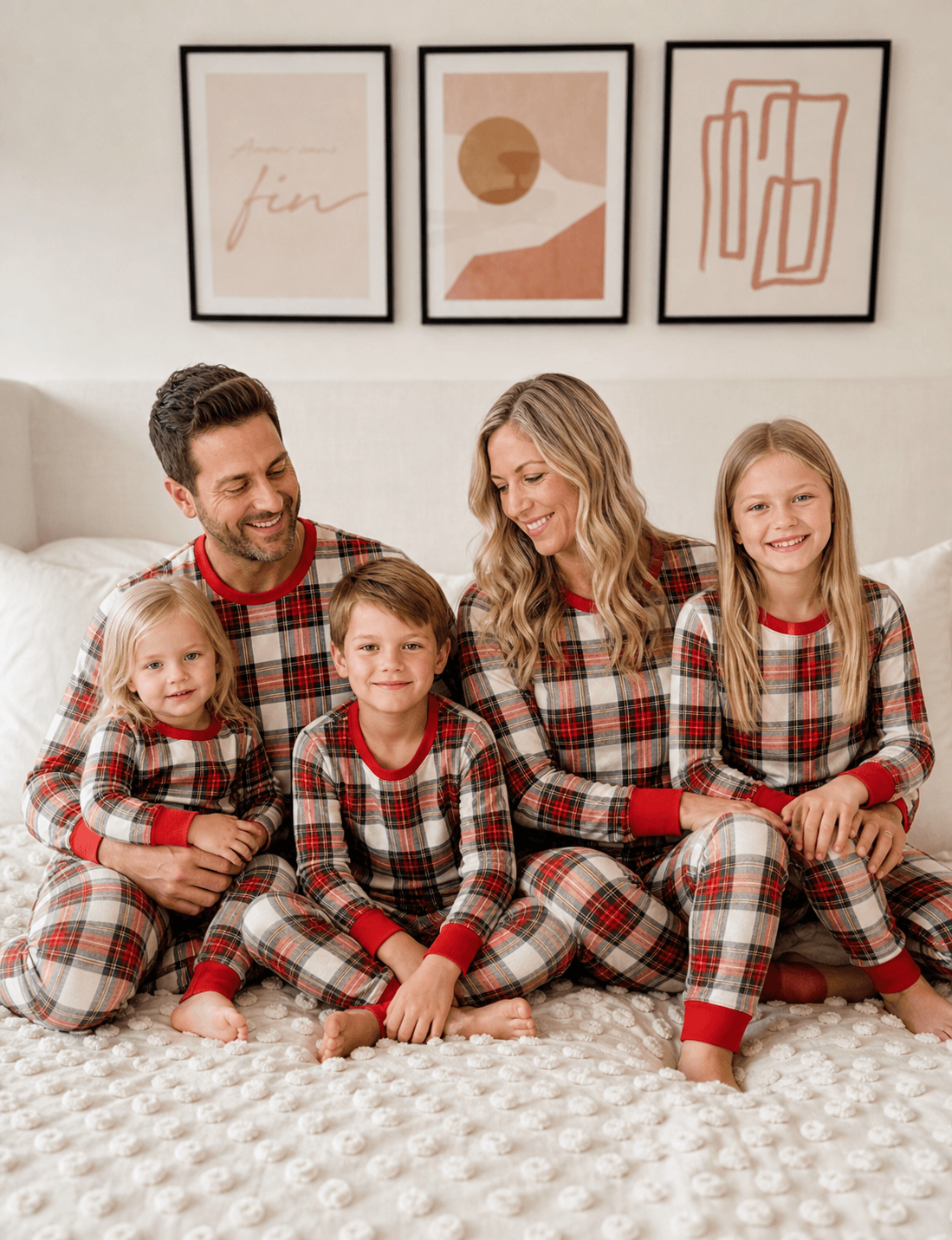 Parents and three children smiling together on a bed in red and white plaid Christmas pajamas with red cuffs.