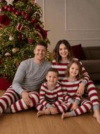 Family sitting on a wooden floor by a Christmas tree in red and white striped pajamas, dad in a grey top.