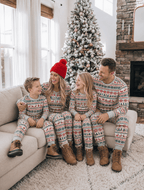 Family in grey Fair Isle Christmas pajamas gathered on a sofa beside a snowy tree and stone fireplace.