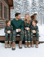 Family holding mugs on snowy cabin steps in green animal-print Christmas pajamas, with dad in a solid green top.