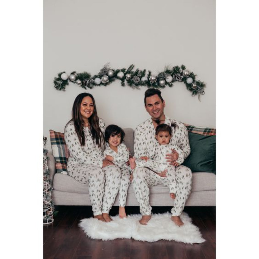 A family of four including a toddler and a baby sitting on a sofa on a fluffy white rug, all modeling matching white loungewear printed with green watercolor pine trees.