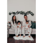 A family of four including a toddler and a baby sitting on a sofa on a fluffy white rug, all modeling matching white loungewear printed with green watercolor pine trees.