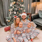 Family of four including parents and twin babies sitting by a decorated Christmas tree wearing matching Fair Isle holiday pajamas.
