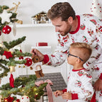 Father and young boy in matching red and white Santa print pajamas hanging a teddy bear ornament on a Christmas tree.