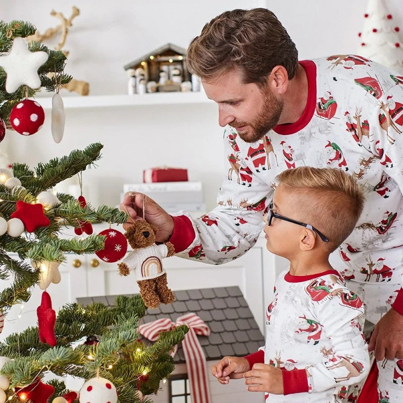 Father and young boy in matching red and white Santa print pajamas hanging a teddy bear ornament on a Christmas tree.