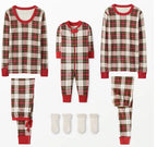 Father and curly-haired son sitting on wooden stools outdoors looking at each other, wearing matching cream, red, and green plaid Christmas pajamas.