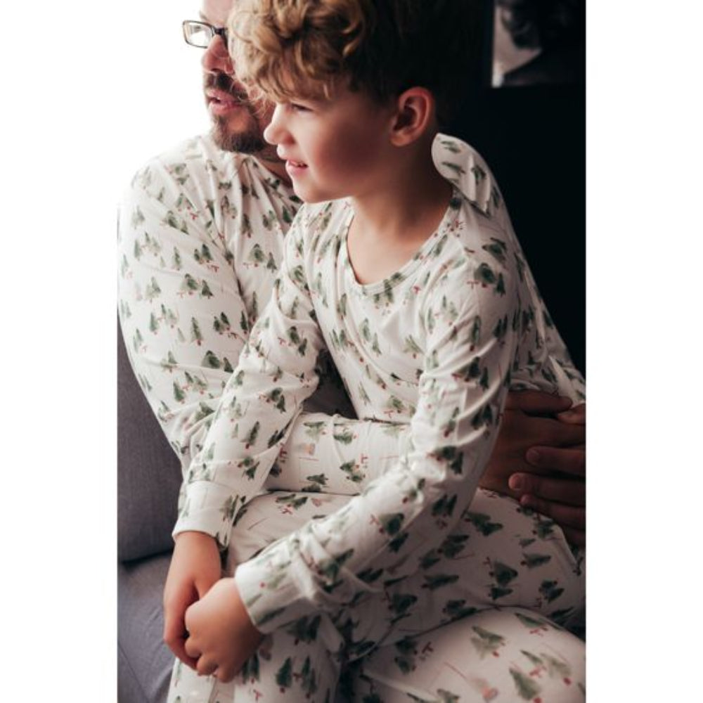 A young boy sitting affectionately on his father's lap, both wearing matching white and green watercolor pine tree print long-sleeve holiday pajamas.