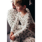 A young boy sitting affectionately on his father's lap, both wearing matching white and green watercolor pine tree print long-sleeve holiday pajamas.