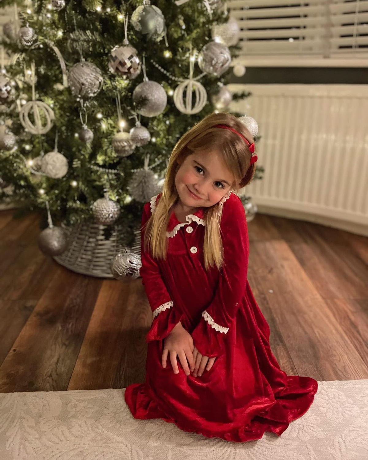 Girl sitting beside a Christmas tree in a red velvet long-sleeve nightgown with white trim.