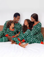 Family of four sitting closely on a bed wearing matching dark green Fair Isle Christmas pajamas with red ribbed cuffs, featuring white snowflake and pine tree patterns.
