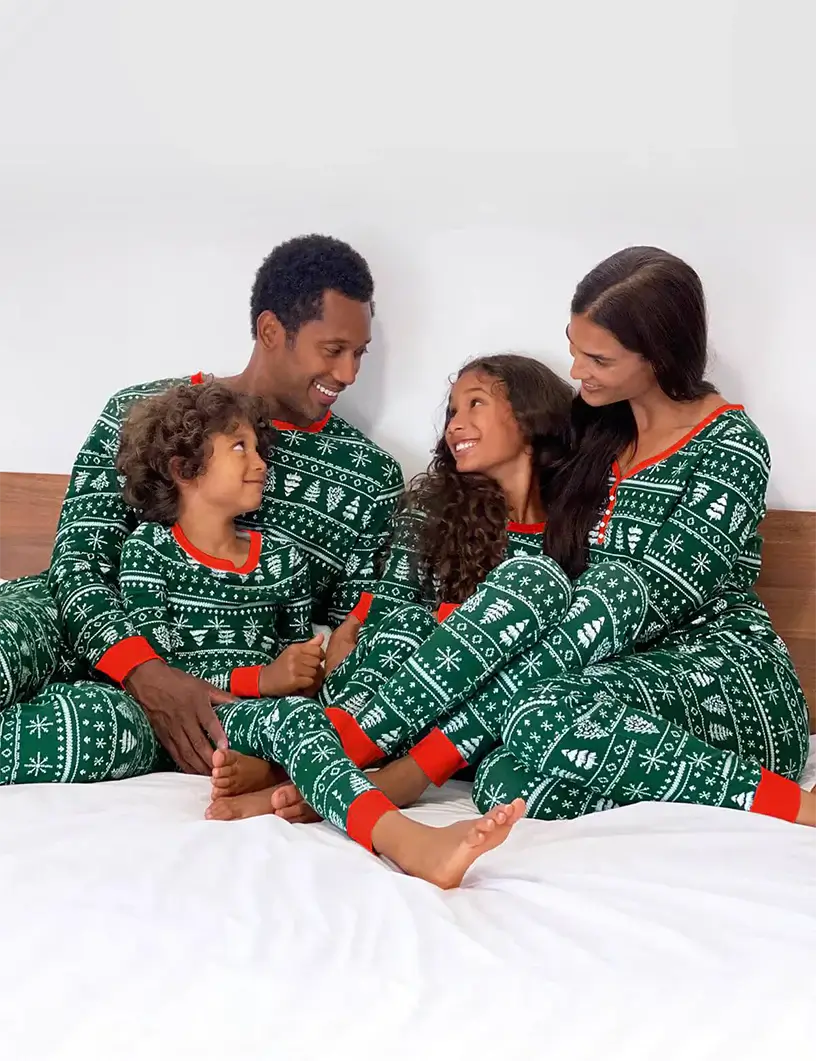 Family of four sitting closely on a bed wearing matching dark green Fair Isle Christmas pajamas with red ribbed cuffs, featuring white snowflake and pine tree patterns.