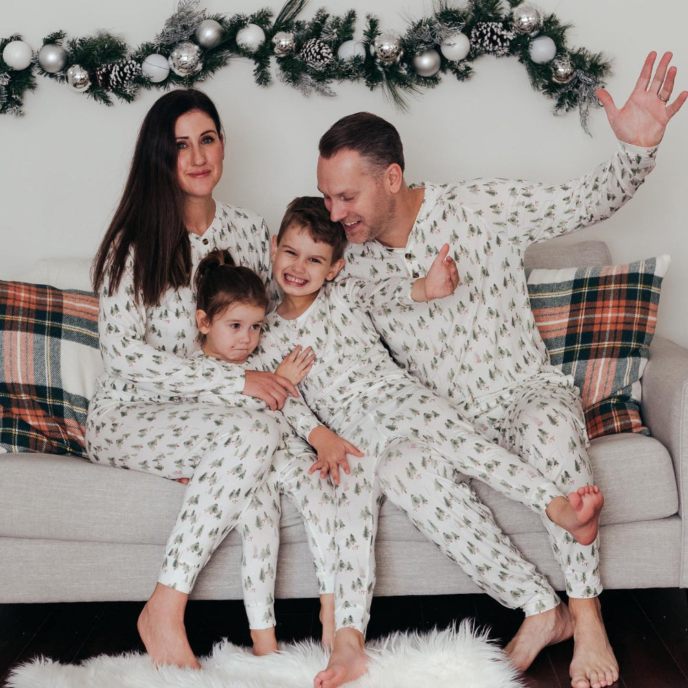 A cheerful family of four sitting on a sofa in matching white two-piece Christmas pajamas with green watercolor tree pattern, with the dad raising his arm in excitement.