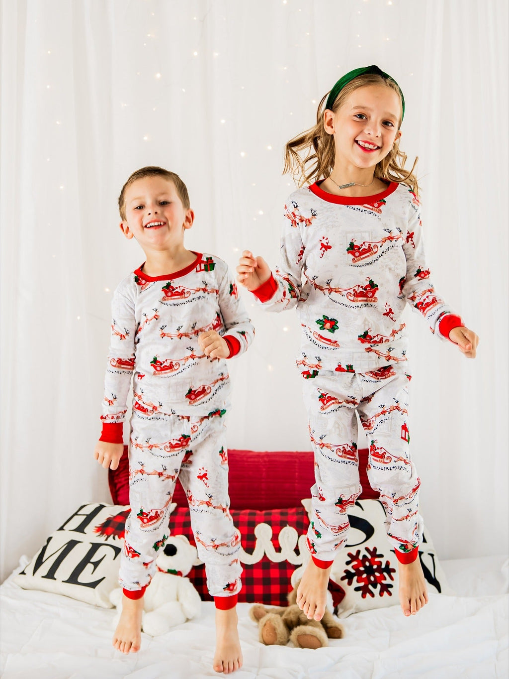 Two happy young kids jumping on a bed in matching white and red Santa print holiday pajamas.
