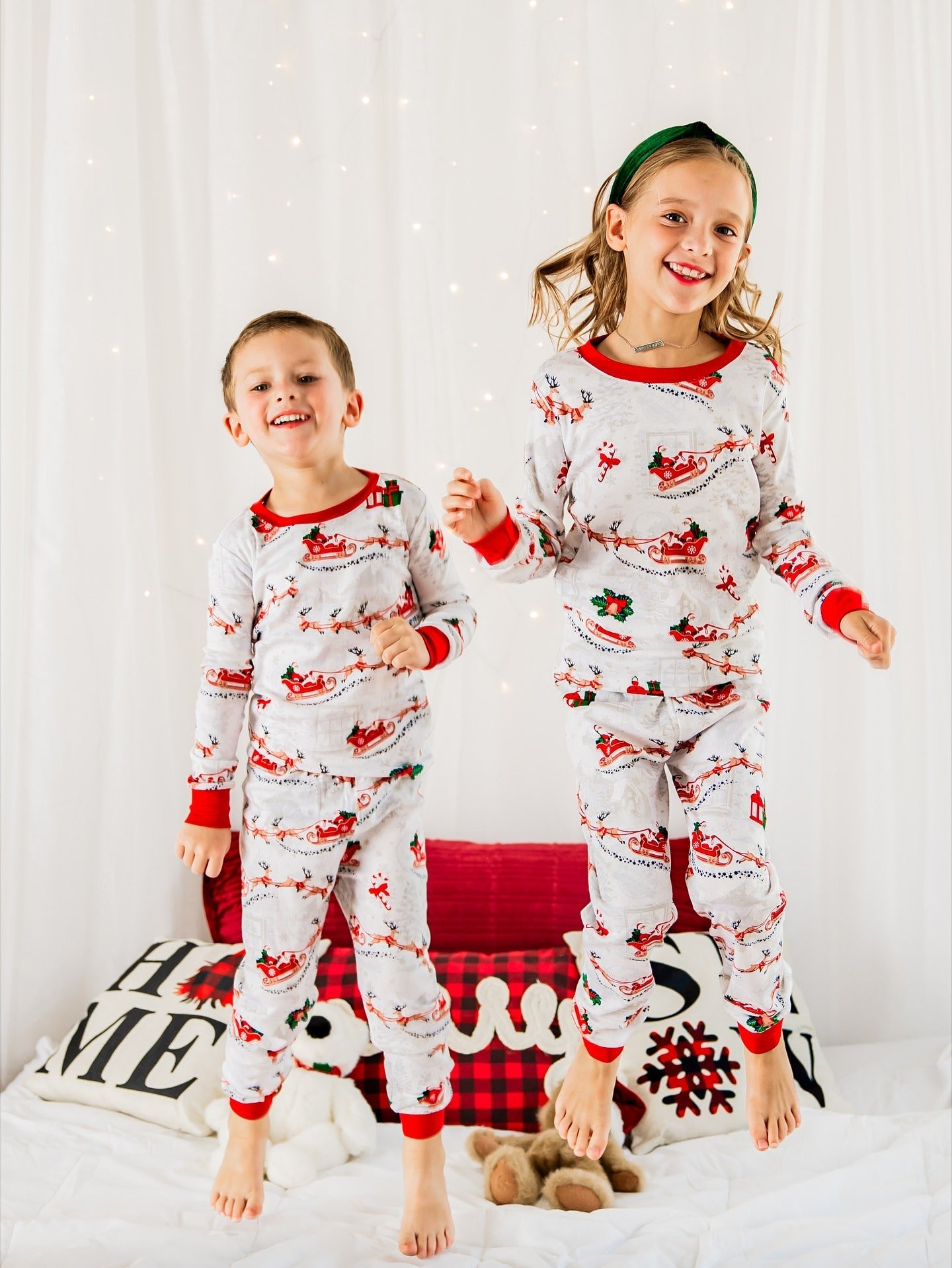Two happy young kids jumping on a bed in matching white and red Santa print holiday pajamas.