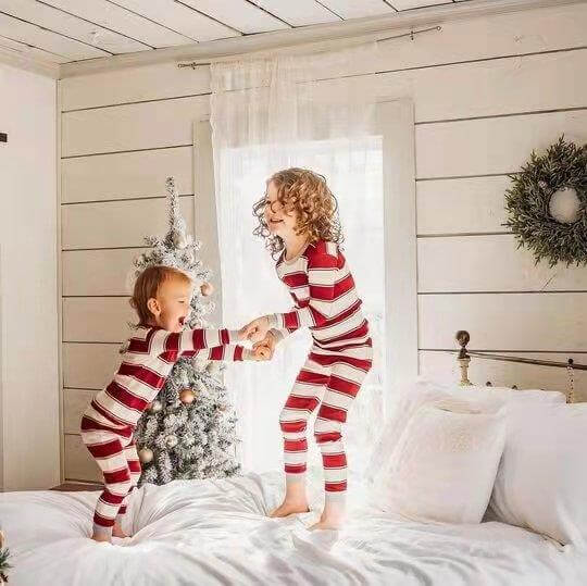 Two young kids holding hands and playing on a white bed wearing snug-fit red and white candy cane striped Christmas pajamas.