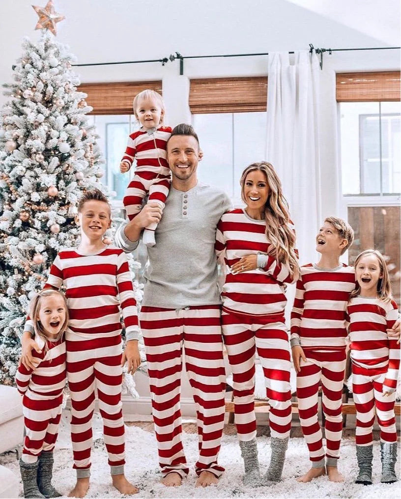 Large family with five kids smiling in front of a frosted Christmas tree, all wearing matching red and white striped holiday pajamas with dad in a grey henley top.
