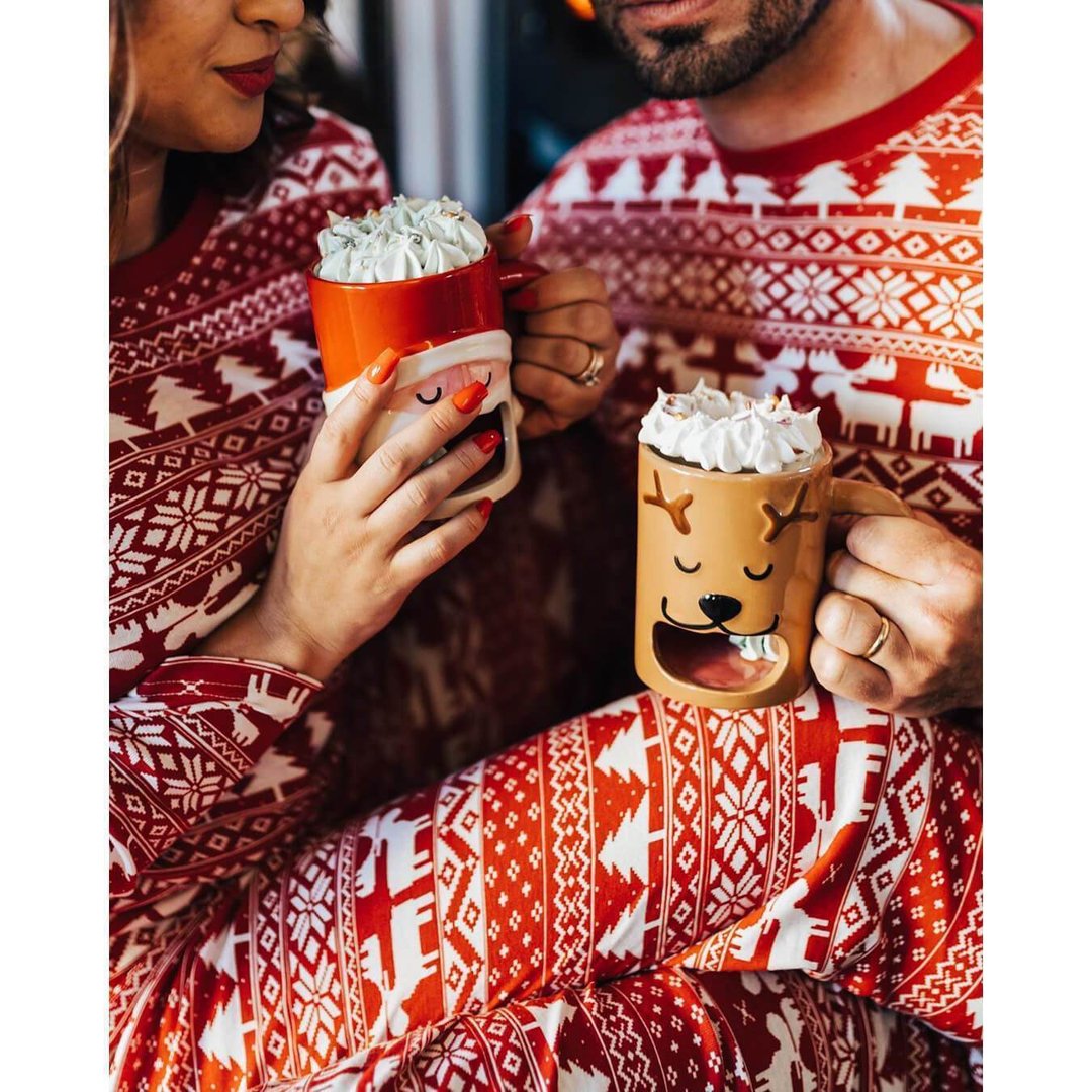 Close-up shot of a couple wearing matching red Fair Isle pajamas, holding festive ceramic Santa and reindeer mugs filled with hot chocolate and whipped cream.