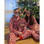 Parents and toddler wearing funny Santa and Christmas tree party glasses while sitting in matching red Fair Isle holiday pajamas in front of a Christmas tree.