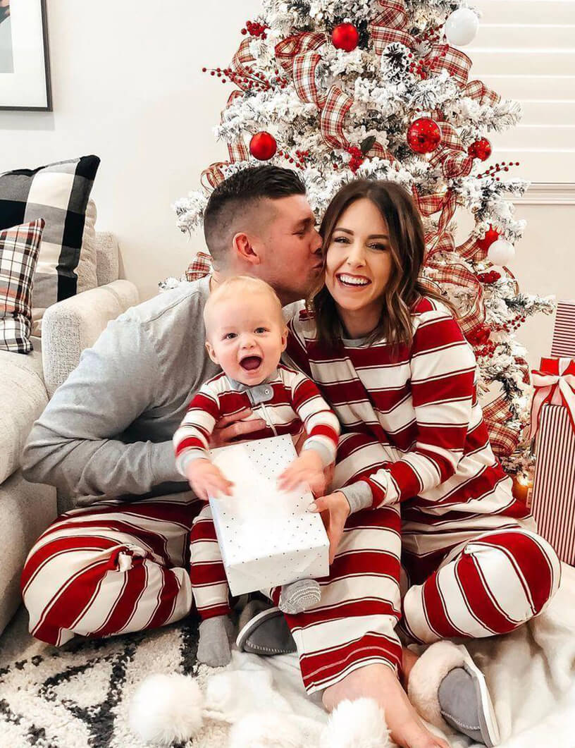 Parents kissing baby while opening Christmas presents under a frosted holiday tree wearing matching red and white striped organic sleepwear.