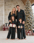 Family of four in matching dark green tartan pajamas holding gifts beside a snow-covered Christmas tree and a stone wall.