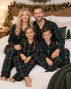 Family of four in matching dark green tartan pajamas seated on a bed with Christmas lights.