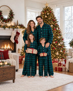Family of four in matching green and black plaid pajamas holding gifts beside a decorated Christmas tree.