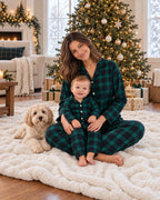 Mother and baby wearing matching green and black plaid pajamas, seated beside a Christmas tree with a cream-colored dog.