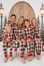 Family seated in red, white and black tartan Christmas pajamas against a snowy arched-door backdrop.