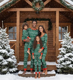 Smiling family standing on a snowy log cabin porch wearing matching green and red Fair Isle Christmas pajamas during winter snowfall.