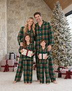 Family of four in matching light green tartan pajamas holding gifts beside a snow-covered Christmas tree and a stone wall.
