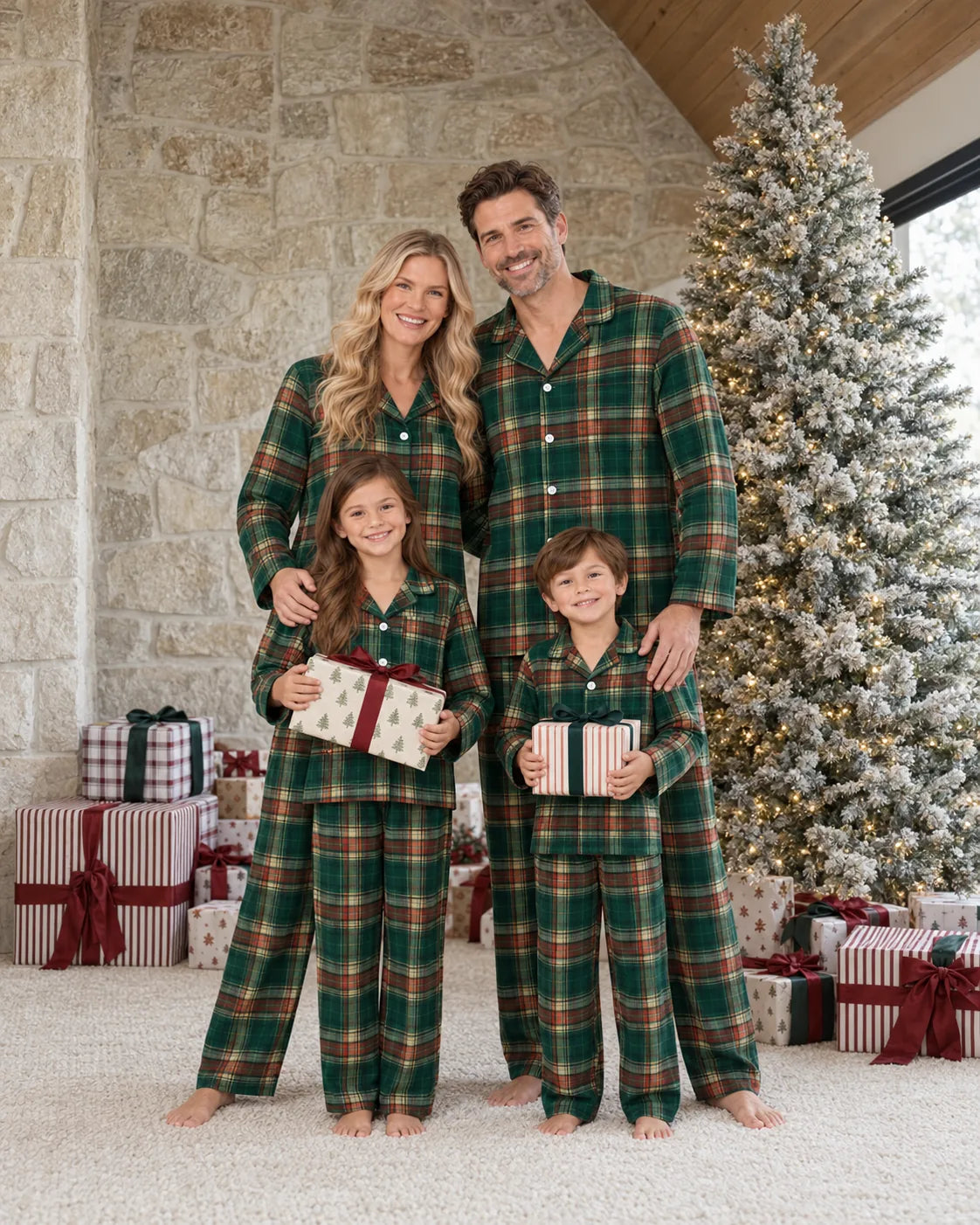 Family of four in matching light green tartan pajamas holding gifts beside a snow-covered Christmas tree and a stone wall.