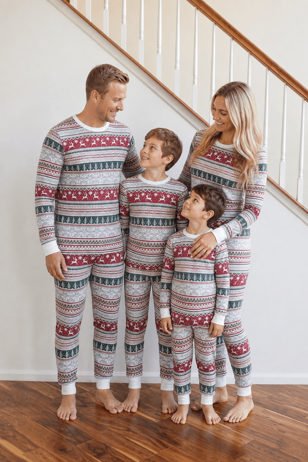 Parents and two boys beside a staircase in light grey Fair Isle Christmas pajamas with red reindeer and green trees.