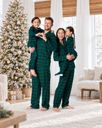 Family wearing forest green plaid matching Christmas pajamas beside a flocked Christmas tree in a bright living room.