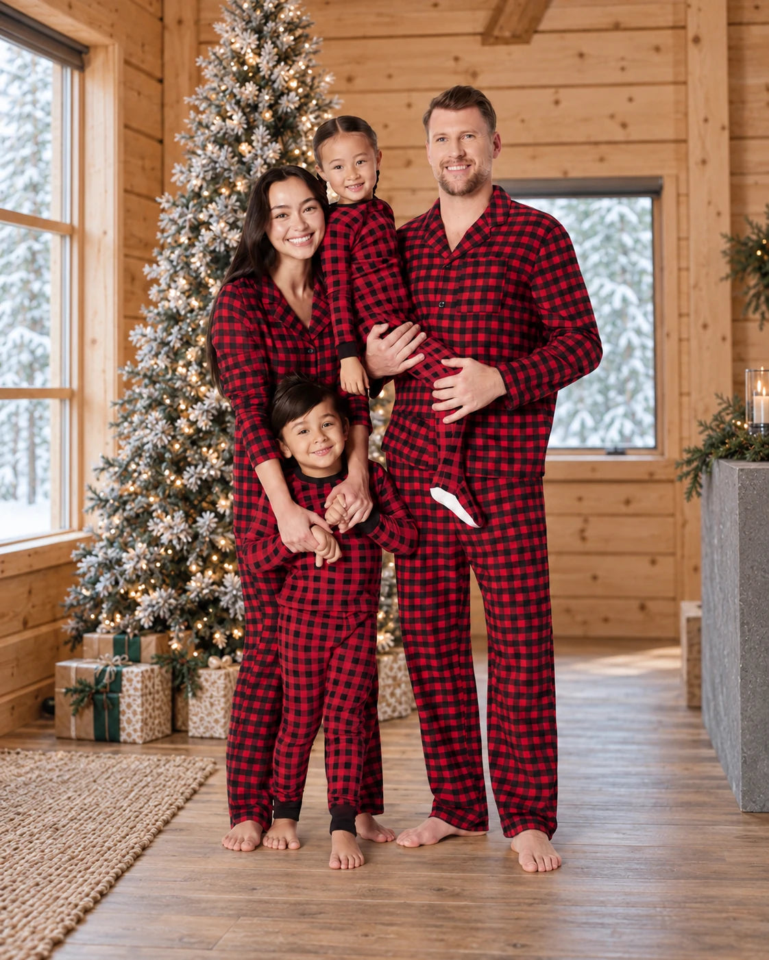 Family wearing red and black plaid matching Christmas pajamas in a snowy Christmas wood cabin.
