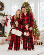 Family of four in matching red and black plaid pajamas holding gifts beside a decorated Christmas tree.