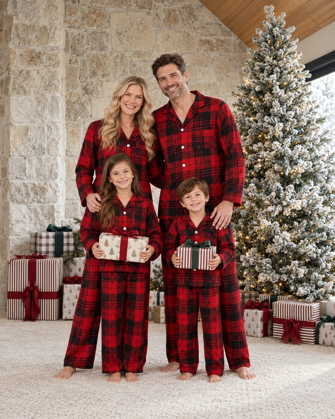Family of four in matching red and black plaid pajamas holding gifts beside a snow-covered Christmas tree and a stone wall.