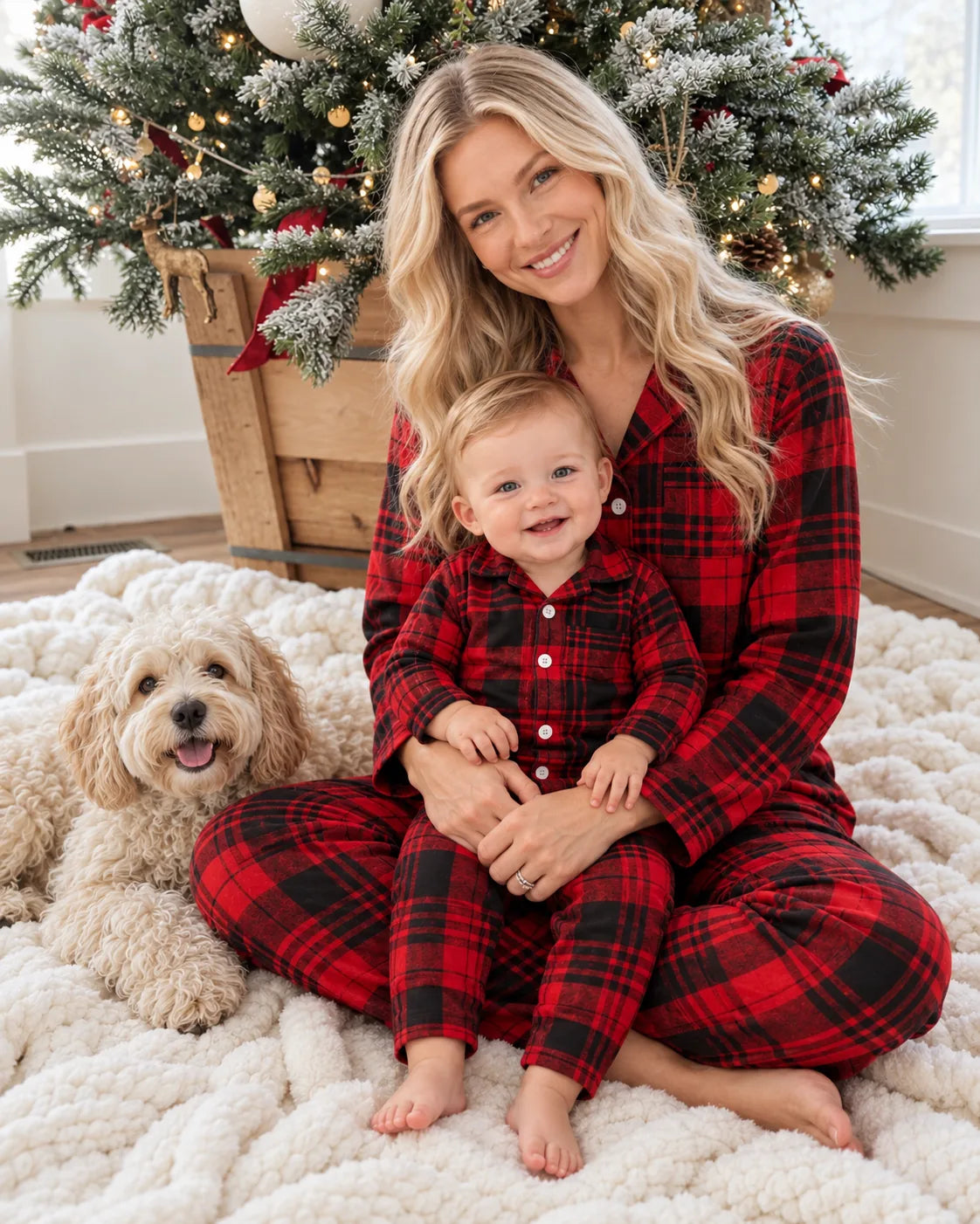 Mother and baby wearing matching red and black plaid pajamas, seated beside a Christmas tree with a cream-colored dog.