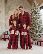 Family of four in matching red and blue plaid pajamas holding gifts beside a snow-covered Christmas tree and a stone wall.