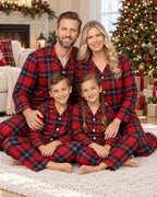 Family of four in matching red and blue plaid pajamas seated on a rug beside a Christmas tree.