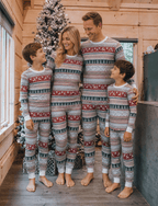 Family in a wood-paneled room wearing matching light grey Fair Isle Christmas pajamas beside a snowy tree.